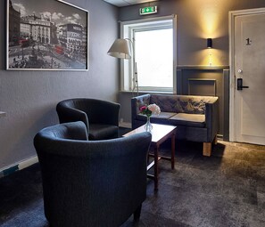 A hotel room with a seating area featuring two armchairs and a sofa, a small table with a vase, a fireplace, and a framed cityscape picture on the wall. - Saga Hotell AB (Borlange)