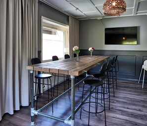 A conference room with a wooden table, metal bar stools, a large window with curtains, a mounted flat-screen TV, and a decorative hanging light fixture. - Saga Hotell AB (Borlange)
