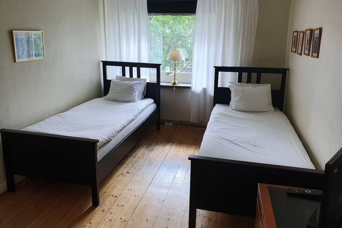 Une chambre avec deux lits simples, une petite table, une lampe et une fenêtre avec des rideaux.