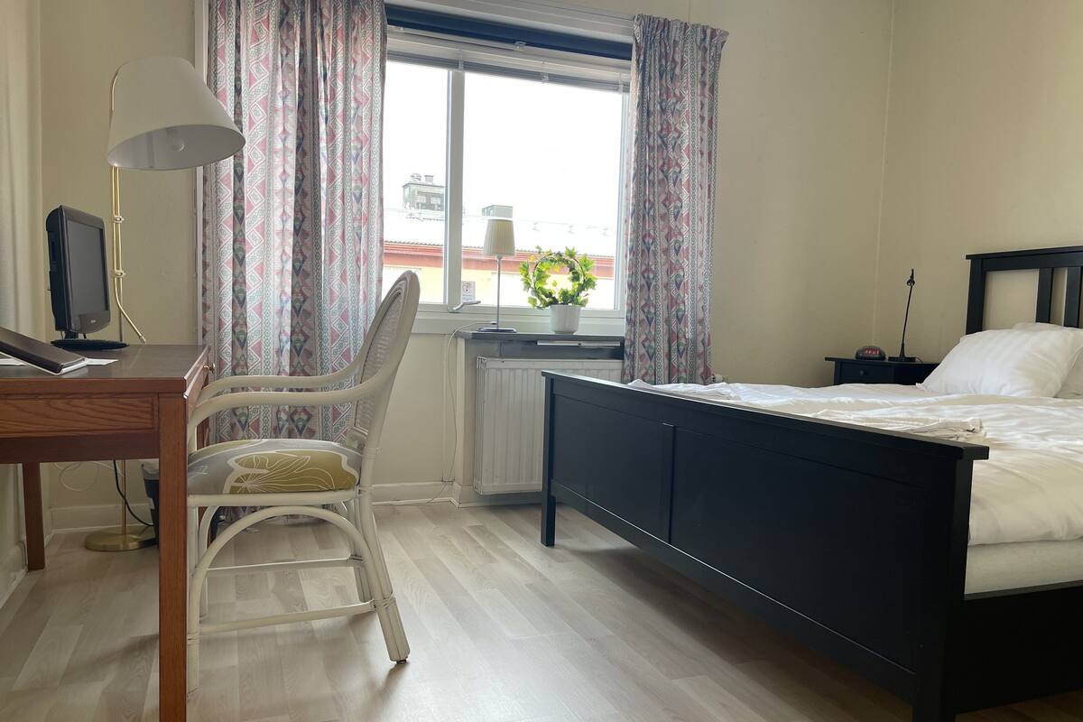 Une chambre à coucher avec un lit, un bureau avec un ordinateur, une chaise, une fenêtre avec des rideaux et un radiateur.