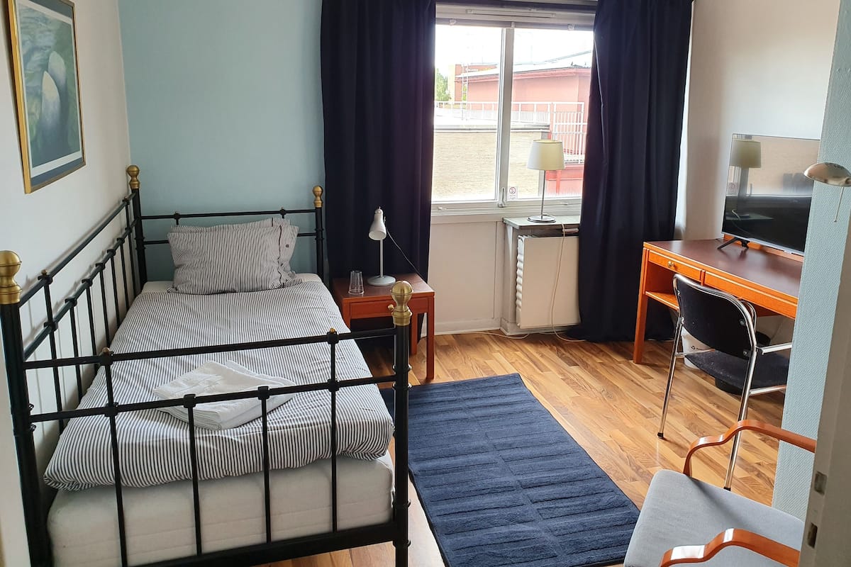Une pièce comprenant un lit, un bureau, une chaise, un radiateur et une fenêtre avec des rideaux.