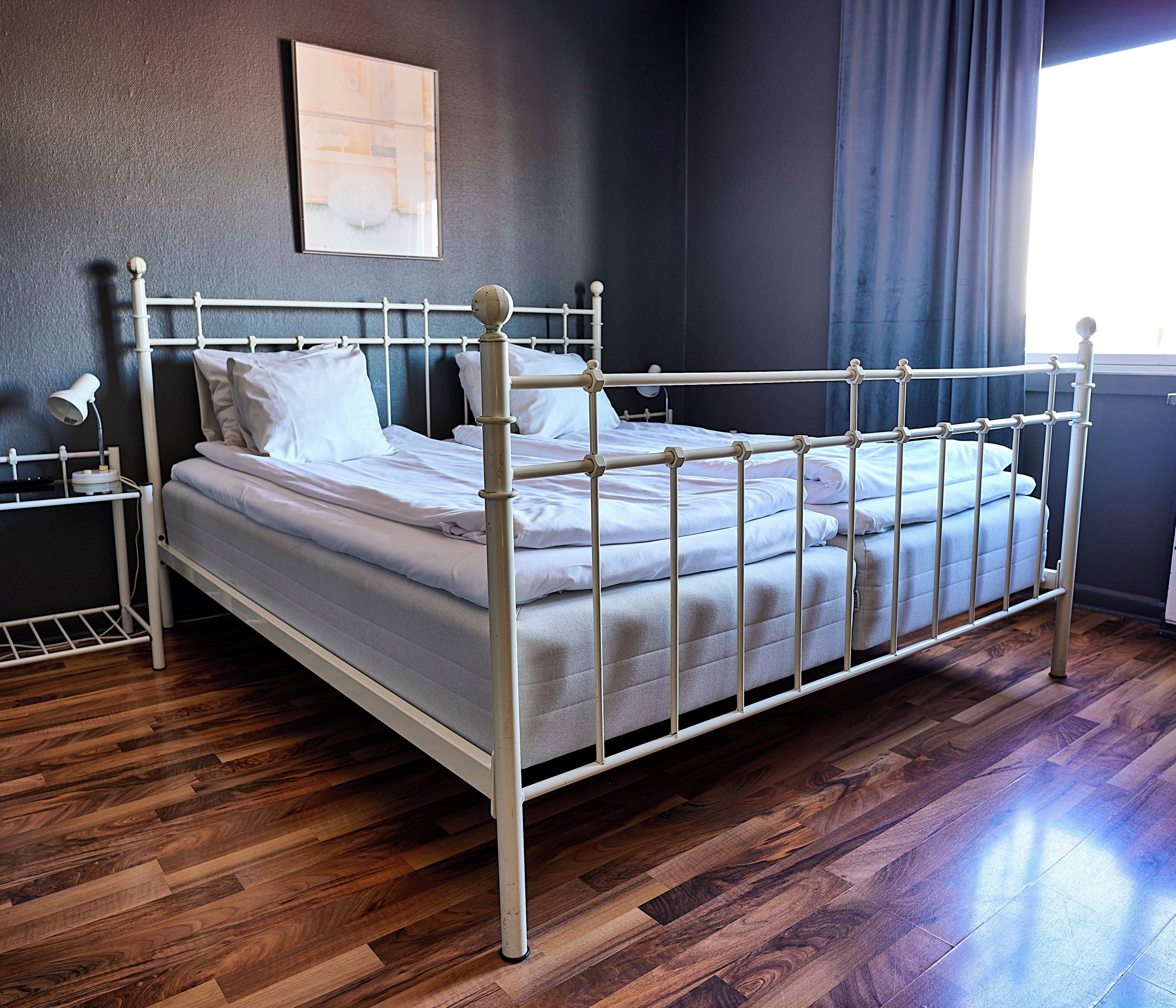 A single bed with a metal frame and white bedding in a room with wooden flooring and a dark wall.