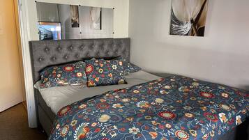 Un dormitorio con cabecera tapizada en gris, colcha con estampado floral y una sola mesita de noche.