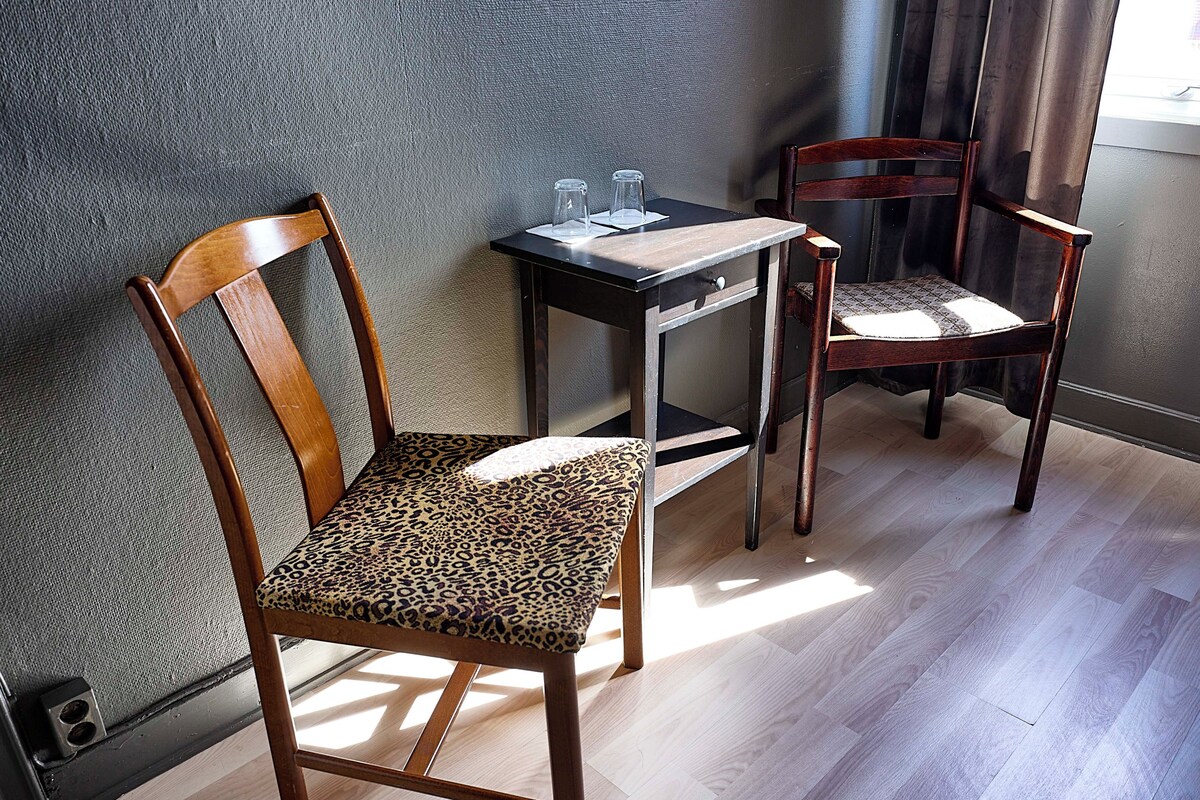 A room with a wooden chair with a leopard print cushion, a small table with two glasses, and a second wooden chair with a plain cushion.