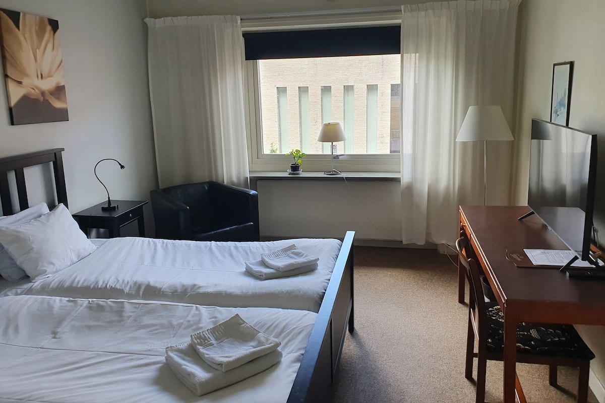 Une chambre d’hôtel avec deux lits, un bureau, une chaise, une télévision et une fenêtre avec des rideaux.