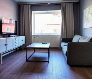 A living room with a television, a sofa, a table, and a window with curtains. - Saga Hotell AB (Borlange)