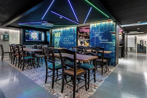 A modern dining area with wooden tables and chairs, a patterned rug, and a wall with a digital screen displaying room information. - Time Hostel NOVA (Jyväskylä)