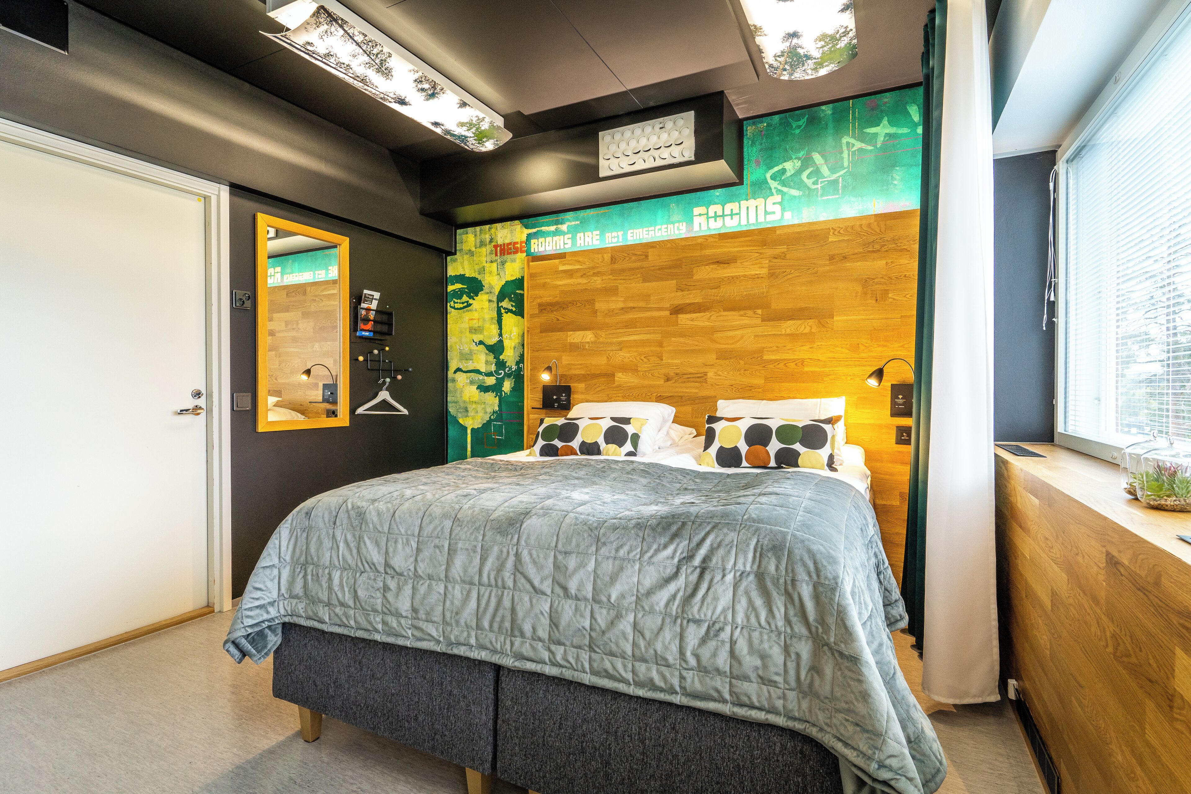 A modern bedroom with a large bed, a wooden headboard, a mirror, and a wall with a green and yellow mural.