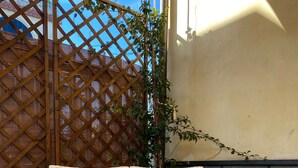 Un sofá de mimbre con un cojín, una mesa blanca con una botella y vasos, y una estructura de celosía de madera.