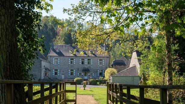 A wooden bridge leading to a stone building with multiple windows and a chimney, surrounded by trees and greenery.