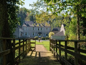 Eine Holzbrücke führt zu einem Steinhaus mit mehreren Fenstern und einem Schornstein, umgeben von Bäumen und Grün.