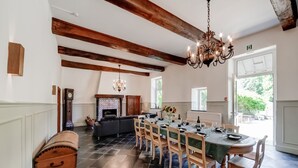 A dining area with a long table set for six, wooden chairs, a chandelier, a fireplace, and a view of the outdoors.