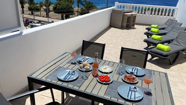 A dining table set with plates, cutlery, and glasses, overlooking a beach with lounge chairs and a view of the ocean. - Stunning front line 2 bedroom apartment. (Tías)