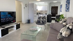 A modern living room with a flat-screen TV, a glass coffee table, a grey sofa, and a dining area with a chair and table. - Stunning front line 2 bedroom apartment. (Tías)