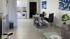 A modern living room with a glass coffee table, a grey sofa, and a dining area with a table and chairs. There is a kitchen in the background with a washing machine and a tiled backsplash. - Stunning front line 2 bedroom apartment. (Tías)