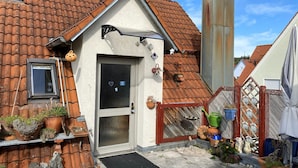 Une maison avec un toit recouvert de tuiles, une porte avec un paillasson noir et des plantes en pot à l’extérieur.