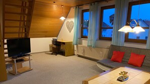 Un salon chaleureux avec une télévision, une table en bois, un canapé avec des coussins rouges et une fenêtre avec des rideaux.