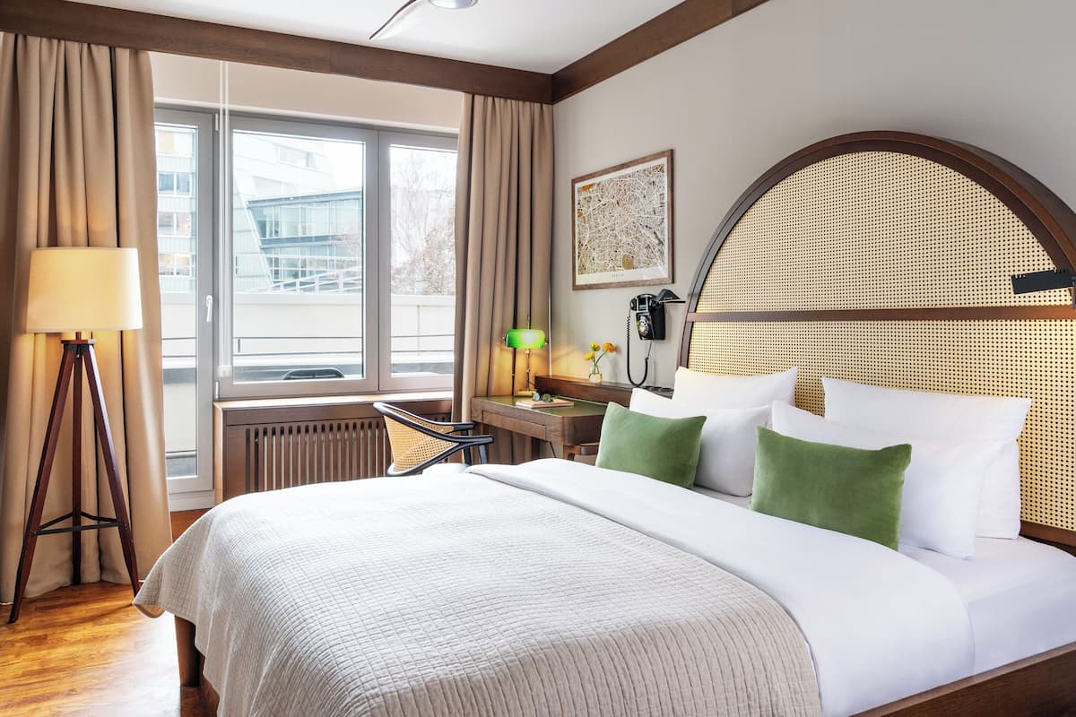 A hotel room with a bed, a desk with a lamp, a telephone, and a window with curtains.