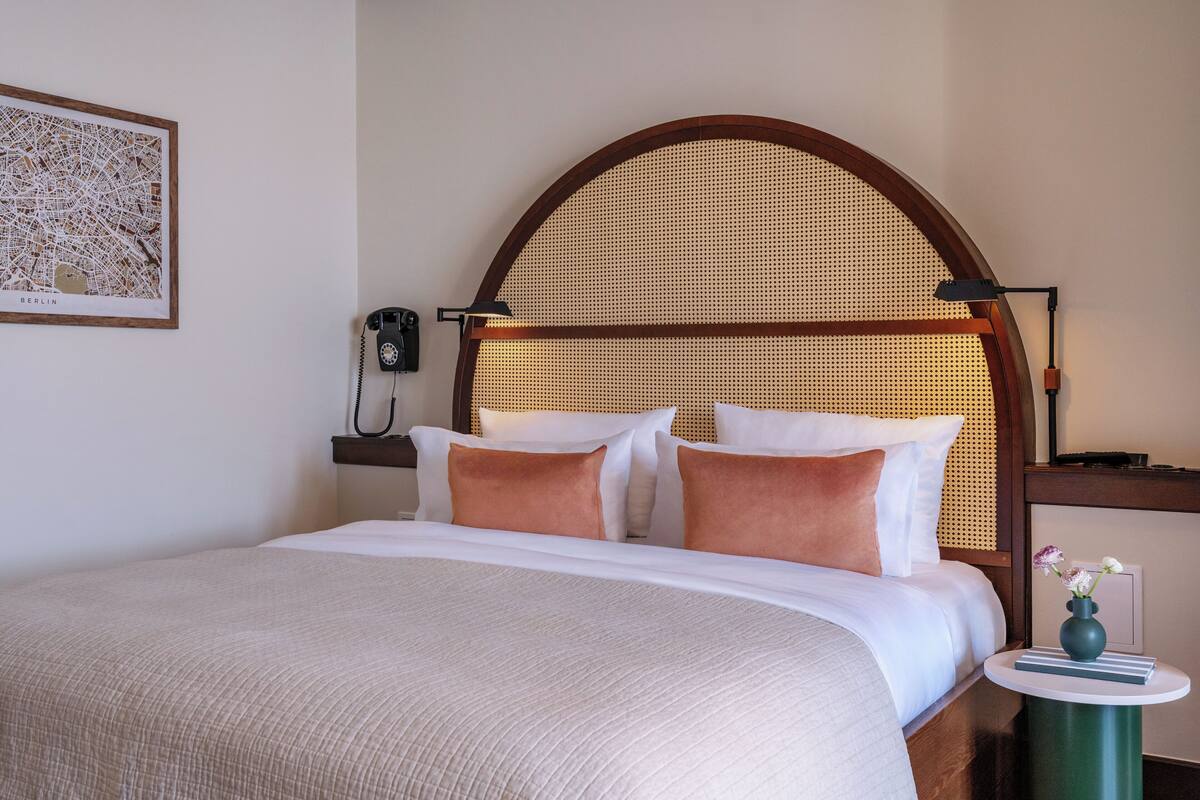 A neatly made bed with a headboard, a bedside telephone, a lamp, and a small table with a vase.