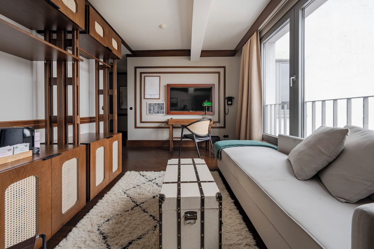 A modern hotel room with a large window, a sofa, a desk with a chair, and a wooden cabinet.