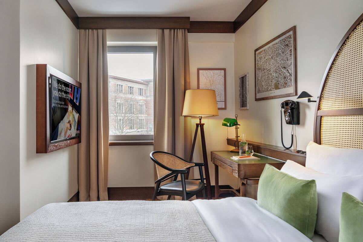 A hotel room with a bed, a desk with a lamp, a chair, a telephone, and a window with curtains.
