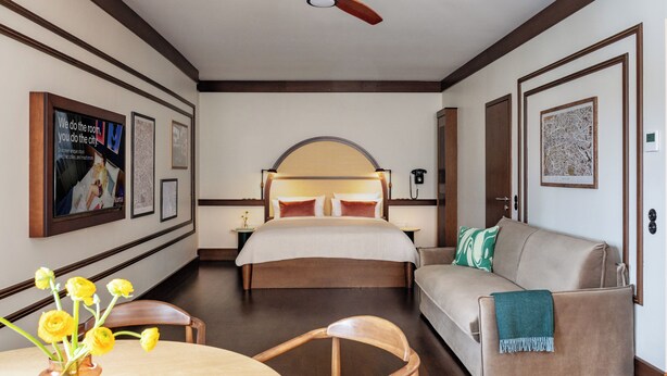 Un cuarto de hotel con una cama, un sofá, una mesa de comedor con sillas, un ventilador de techo, cuadros en las paredes y un jarrón con flores.