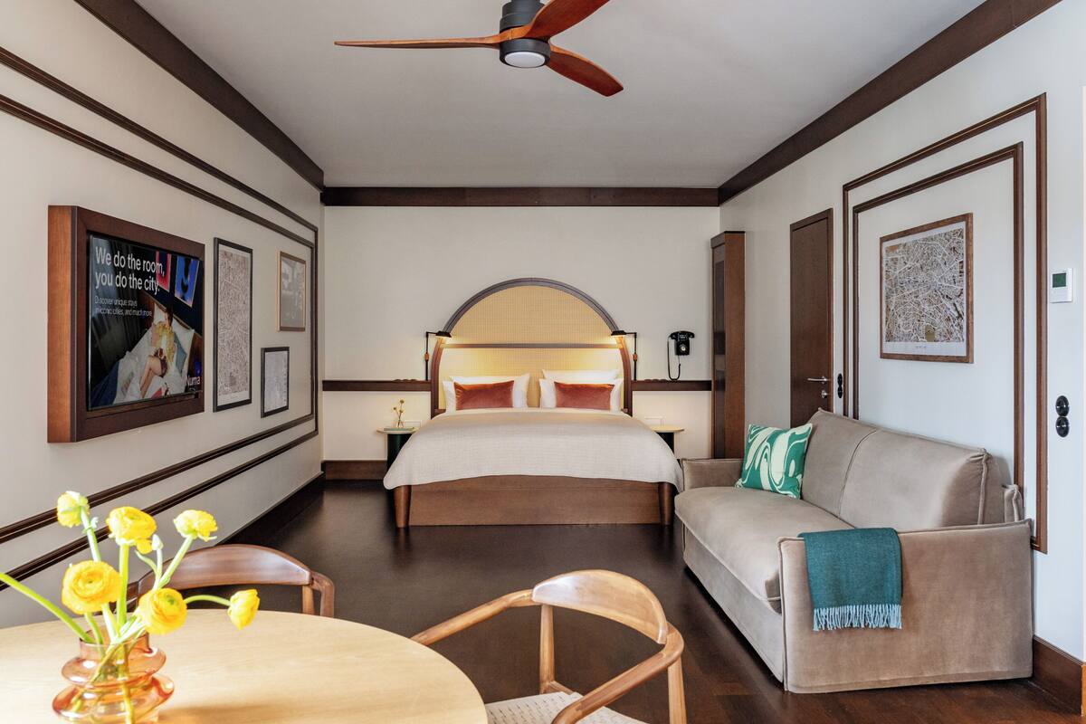A hotel room with a bed, a sofa, a dining table with chairs, a ceiling fan, framed pictures on the walls, and a vase with flowers.