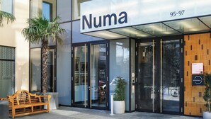 Un edificio moderno con entrada de vidrio, un banco de madera en el exterior y un letrero que dice 'Numa 95-97'.