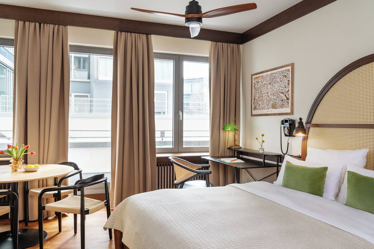 A hotel room with a bed, a desk, a chair, a table with a vase of flowers, a window with curtains, and a ceiling fan.