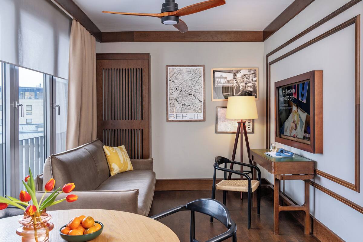 A modern hotel room with a sofa, a dining table with fruit, a lamp, a chair, a desk, and a ceiling fan.