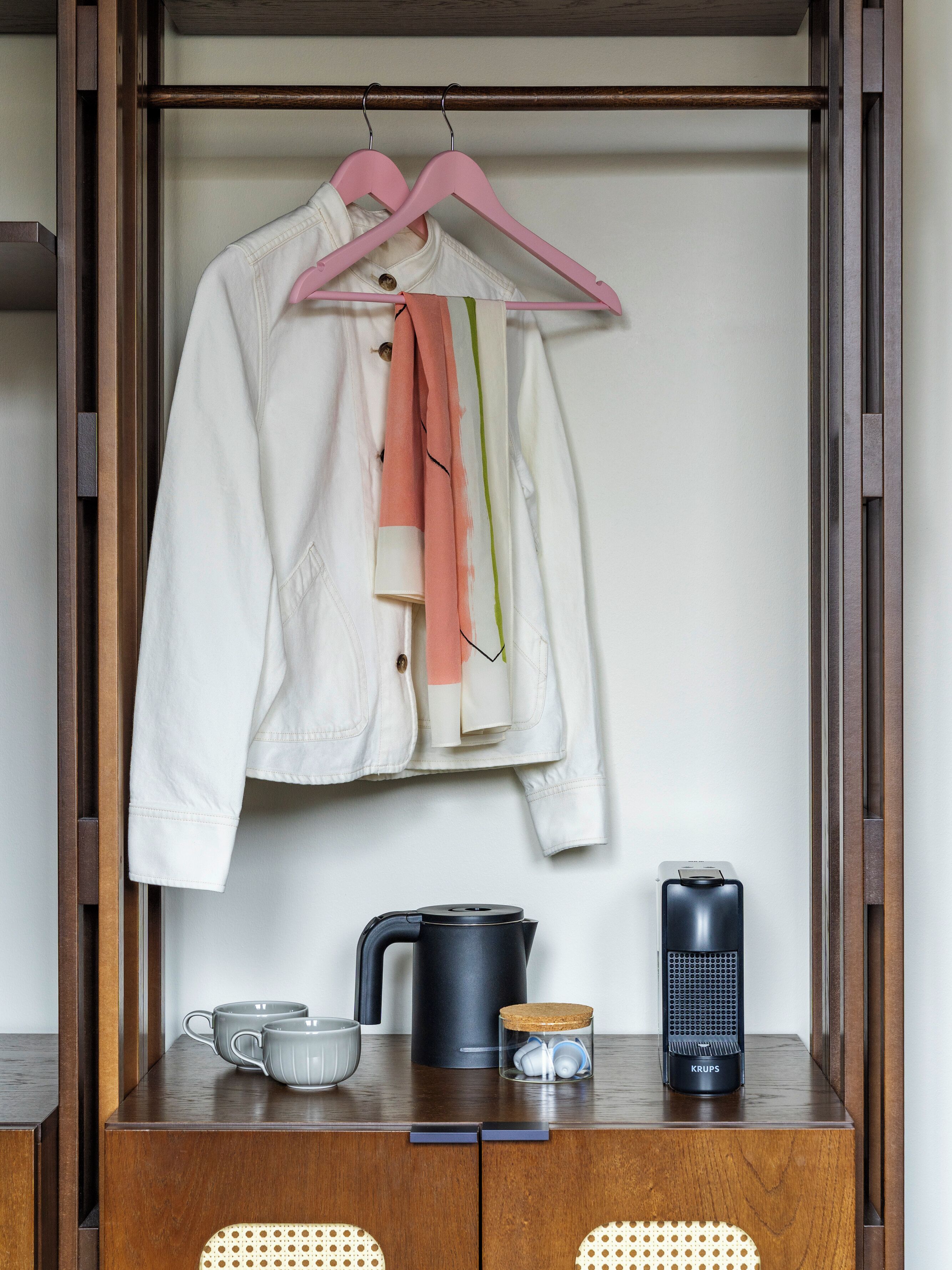 A wooden wardrobe with a hanging rack, a kettle, a coffee maker, and a cup on the lower shelf.