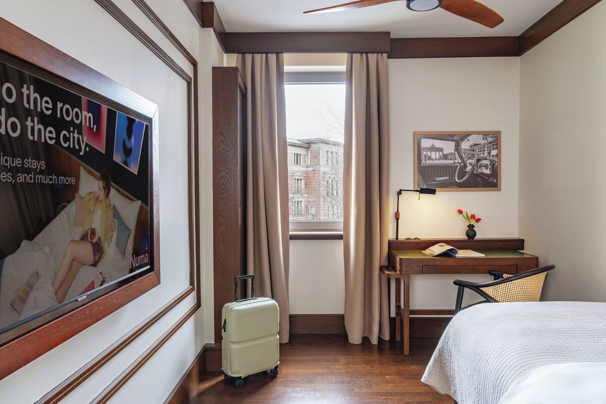 A hotel room with a bed, a desk with a lamp, a suitcase, a window with curtains, and a framed picture on the wall.
