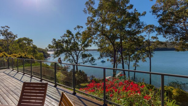 Une terrasse en bois avec une balustrade donnant sur un lac, entourée d’arbres et de fleurs rouges.
