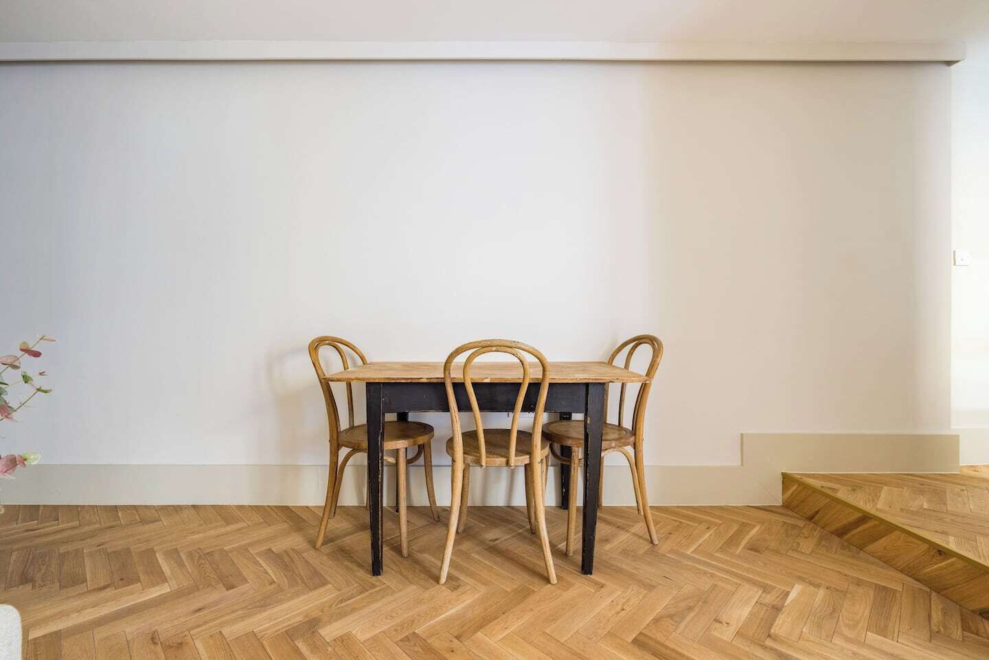 A dining area with a wooden table and chairs, a herringbone-patterned floor, and a white wall.