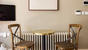 Une pièce avec une table ronde, deux chaises en osier, un radiateur, un grand tableau abstrait et un téléviseur fixé au mur.