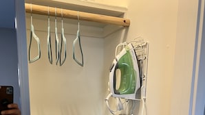A bathroom with a wooden shelf holding multiple hangers, a green iron, and a patterned ironing board.