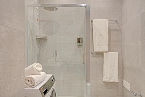 Un baño moderno con ducha envuelta en vidrio, lavadora integrada y dos toallas blancas en un tendedero.
