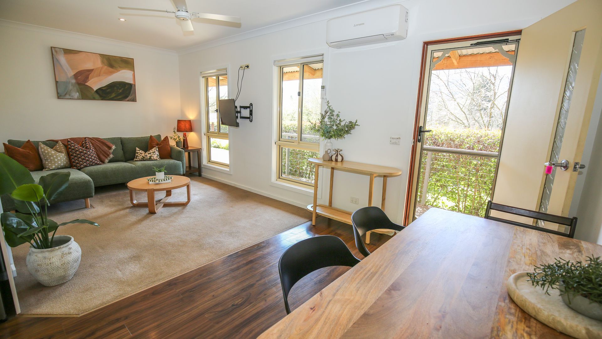 A living room with a green sofa, a wooden coffee table, and a dining table with chairs. There is a ceiling fan, a window with a view, and an air conditioning unit on the wall.