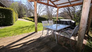 Une terrasse couverte avec une table et des chaises, entourée d’une pelouse bien entretenue et d’arbres.