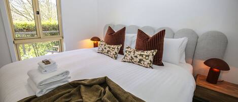 A neatly made bed with pillows, a blanket, and towels, a bedside table with a lamp, and a window with a view of greenery.