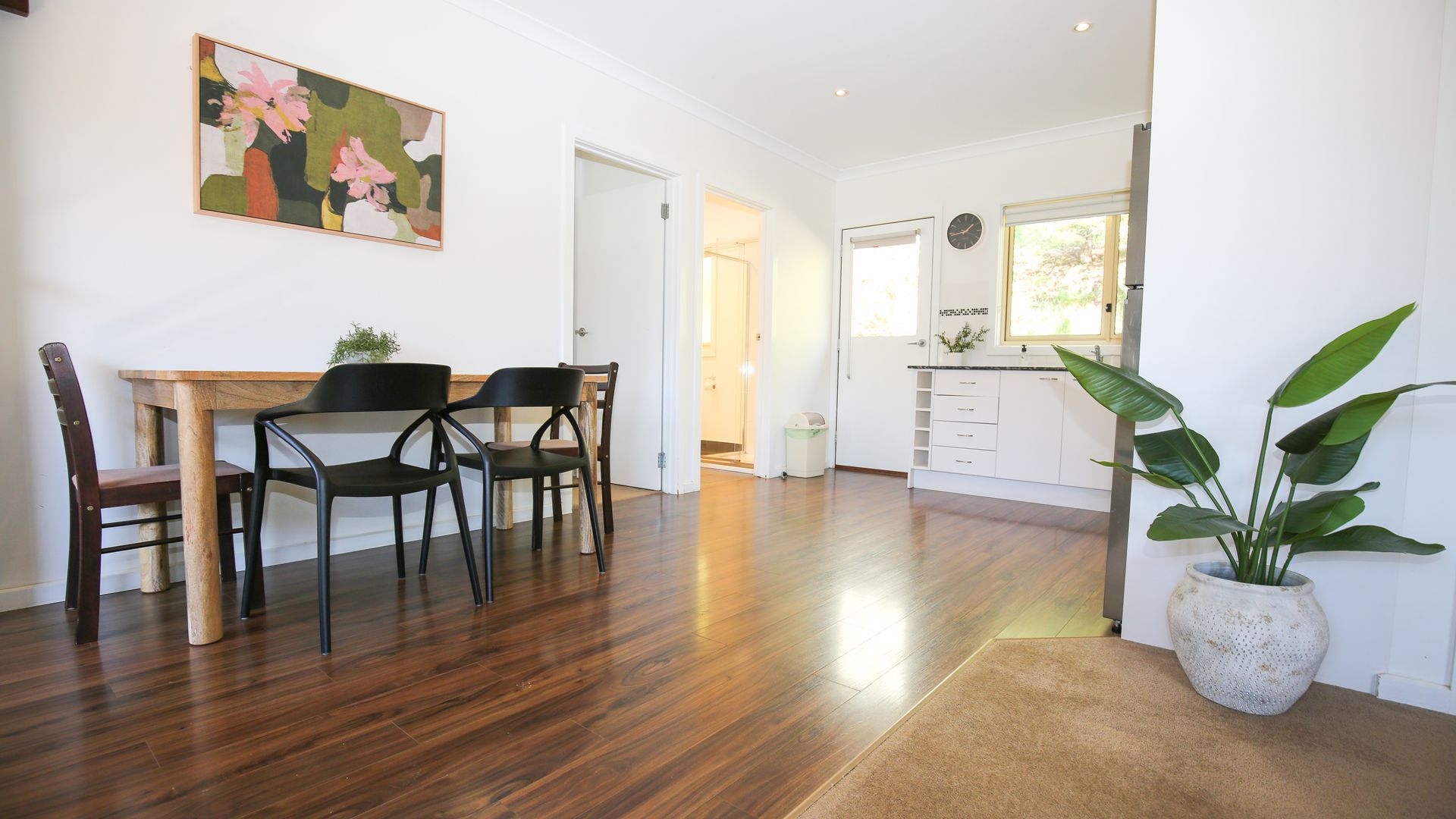 A dining area with a wooden table and chairs, a kitchen with white cabinets, and a potted plant.