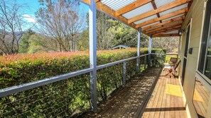 A covered wooden deck with a metal railing, a small table and chairs, and a view of a landscaped garden with bushes and trees.
