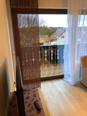 Une pièce avec un plancher en bois, un canapé, une télécommande et une vue sur un balcon agrémenté d’un rideau blanc.