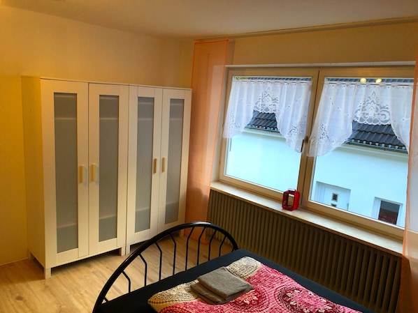 Une pièce avec une fenêtre, un radiateur, une armoire blanche et une table recouverte d’une nappe à motifs rouges et blancs.