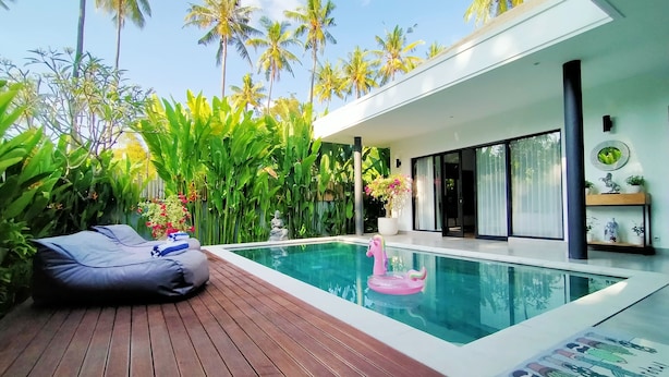 A modern pool area with a wooden deck, a pink flamingo float, and lush tropical plants. - Villa Ataya (Sukasada)