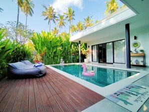 A modern pool area with a wooden deck, a pink flamingo float, and lush tropical plants. - Villa Ataya (Sukasada)