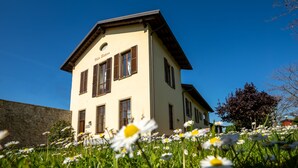 Ein zweistöckiges Gebäude mit einer beige farbenen Fassade und braunen Fensterläden, umgeben von einem Acker voller Gänseblümchen.