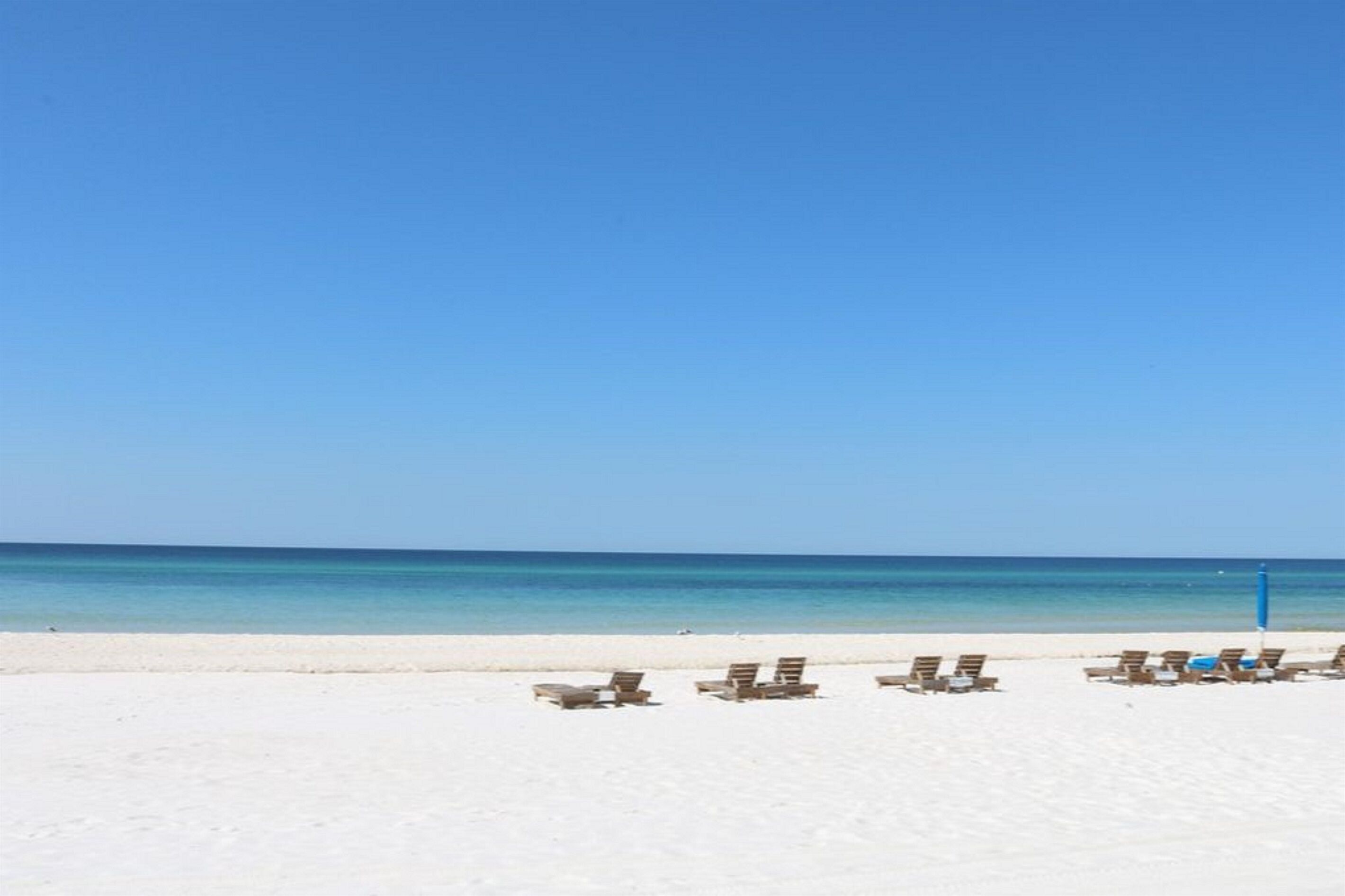 Une plage de sable blanc, sous un ciel bleu azur. Plusieurs chaises longues sont alignées le long du rivage.