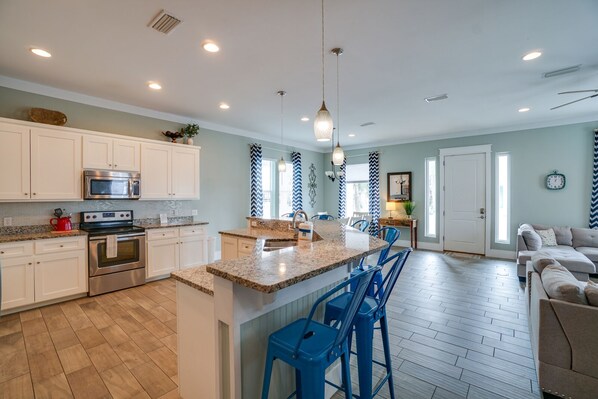A spacious kitchen with white cabinets, a granite countertop, and stainless steel appliances. There is a large island with blue bar stools, and a dining area with a chandelier.