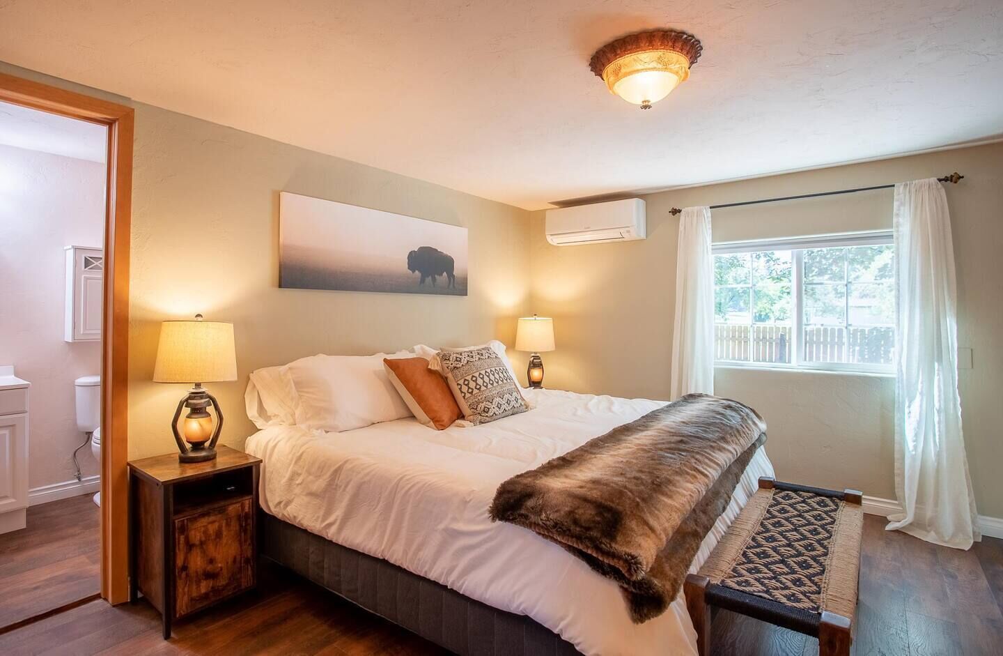 A bedroom with a large bed, two bedside lamps, a wooden nightstand, a bench, a window with curtains, and an air conditioning unit.